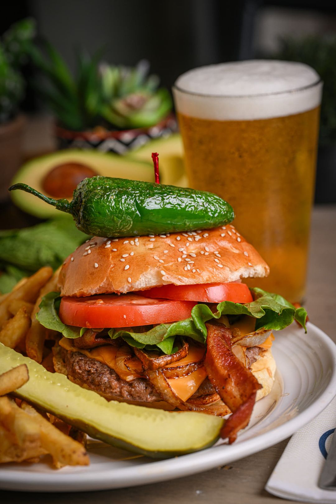 Build-your-own burger stacked with bacon, cheese, grilled onion, lettuce, tomato and a whole jalapeño, fries and a pint of beer behind